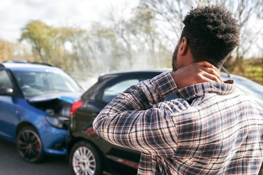Man holds neck after car accident with two vehicles in background, illustrating whiplash injury.