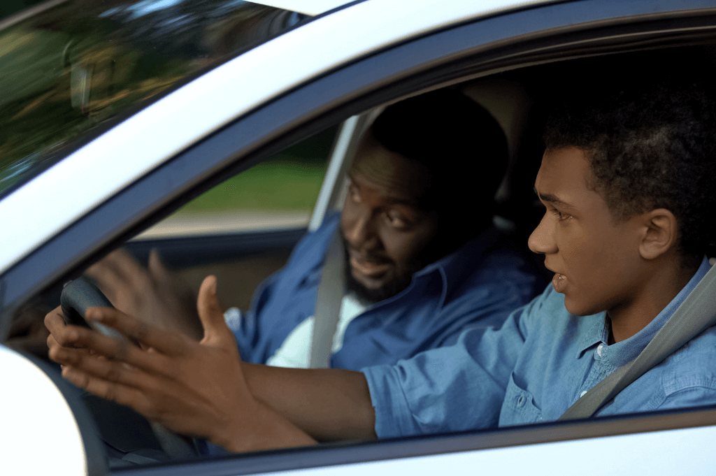 Two individuals in a car; a young male in the front passenger seat gesturing while talking, and a male driver looking on attentively. They appear to be engaged in conversation.