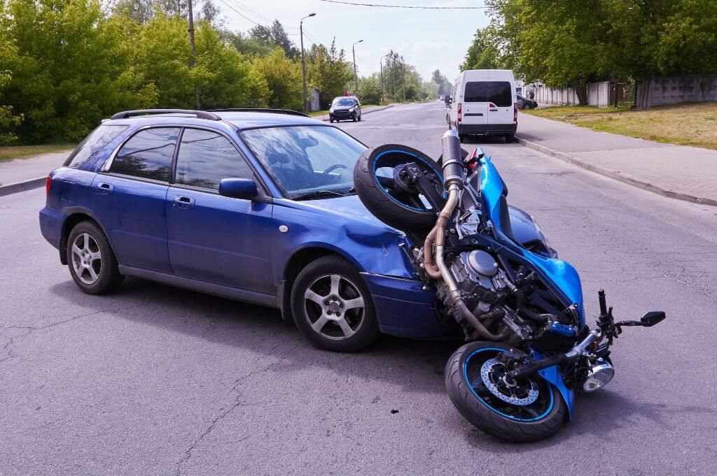 A blue motorcycle lies on its side behind a blue car on a paved road, suggesting a minor traffic accident, with trees and vehicles in the background.