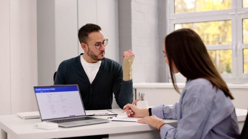 Man with arm cast discusses health insurance claim with a woman in an office setting.