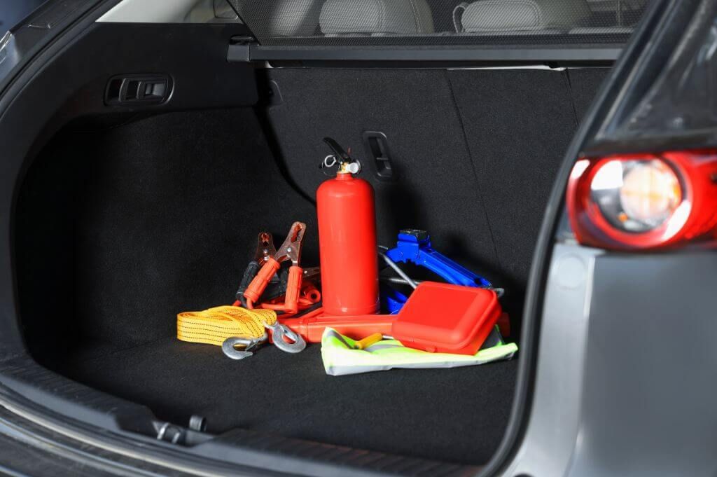 Emergency tools rest inside a car trunk: a red fire extinguisher, jumper cables, tow rope, jack, and first-aid kit, indicating preparedness for vehicular incidents.