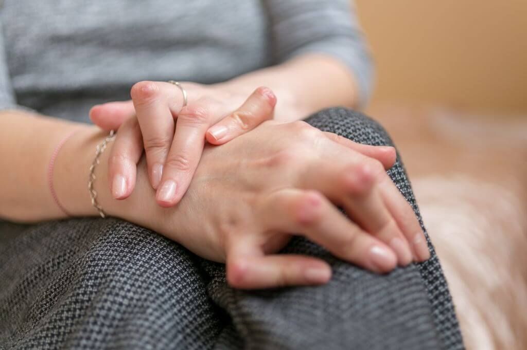Close-up of a person’s clasped hands resting on their lap, suggesting comfort or contemplation, set against a soft-focus indoor backdrop.