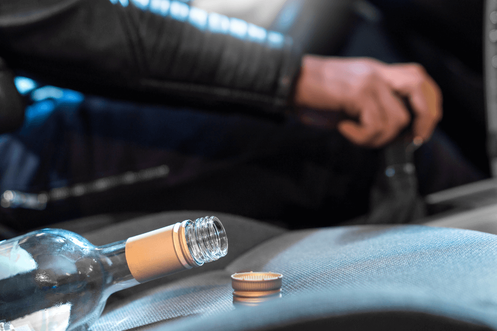 A partially empty glass bottle lies on a car seat, with its cap beside it. A hand grips a gear shift, suggesting the vehicle is in operation.