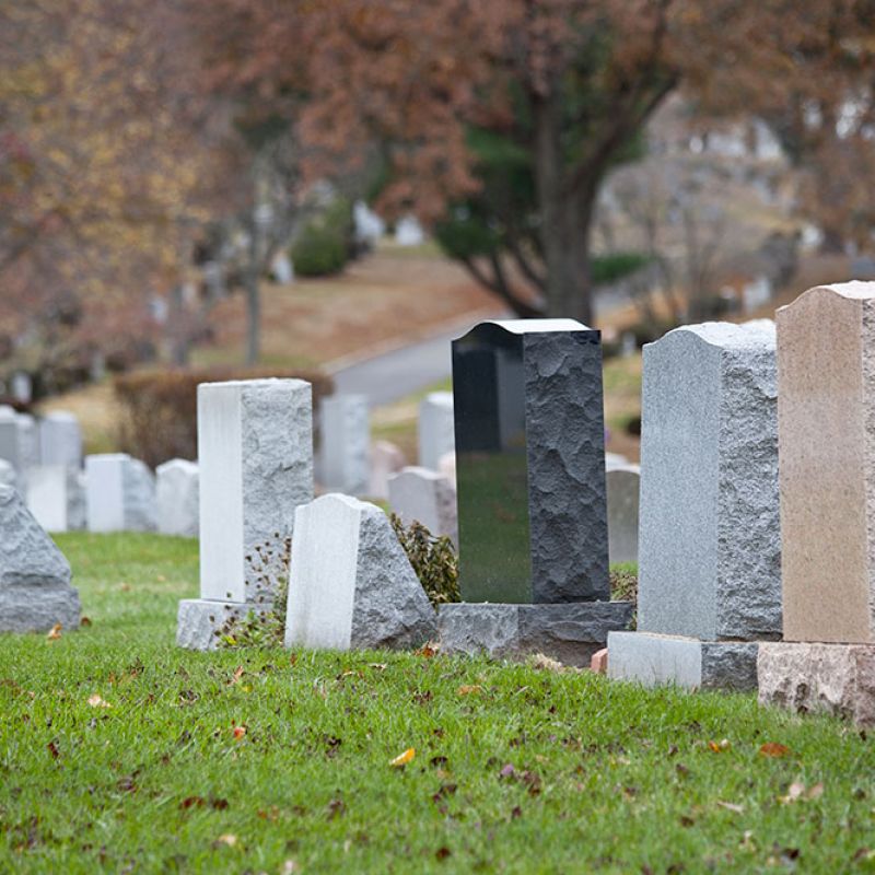 Headstones in a peaceful cemetery setting with green grass and trees in the background during autumn.