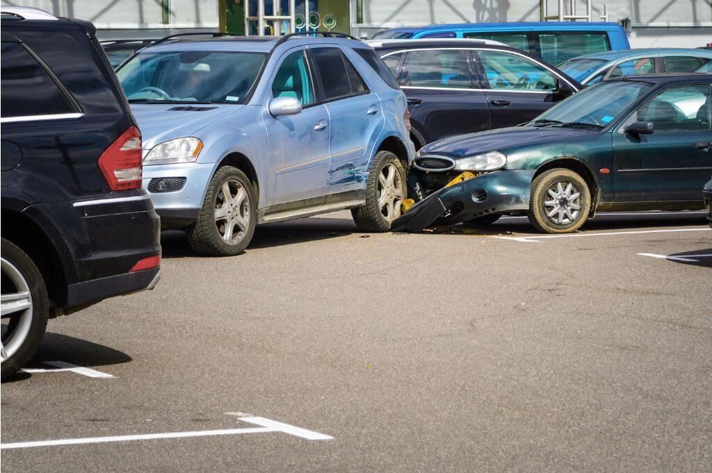 A blue SUV is wedged against a green sedan, which has its front bumper partially dislodged. This accident occurs in a busy parking lot filled with various parked vehicles.