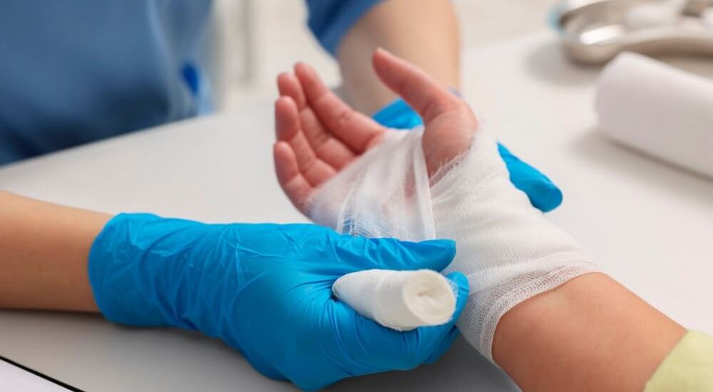 A healthcare professional in blue gloves wraps a bandage around a patient's injured wrist while seated at a table in a clinical setting. The ambiance is sterile and focused on care.