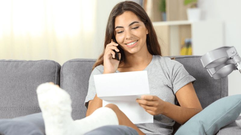 Woman smiling on phone looking at paperwork with leg in cast elevated