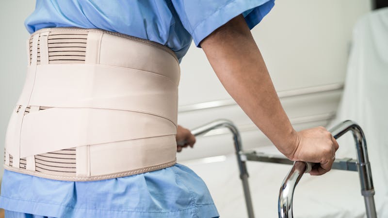 | Matz Injury Law A person wearing a back support belt stands using a walker, likely recuperating, within a clinical or rehabilitation setting.