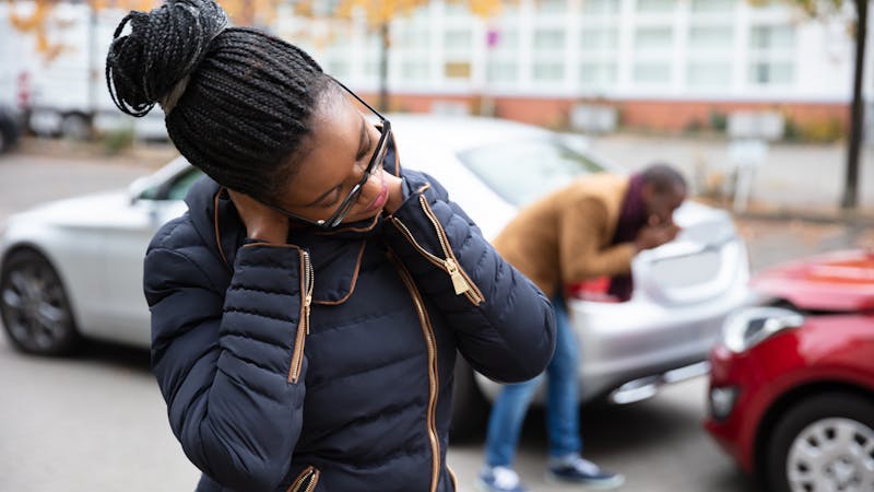A woman standing outside her car holding her neck in pain, possibly suffering from whiplash, while another person inspects the damage to the rear end of her car after an accident.