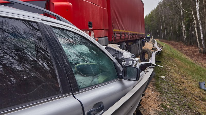 A damaged car with its front end crushed is wedged under a red semi-truck trailer on the side of a road, surrounded by trees.