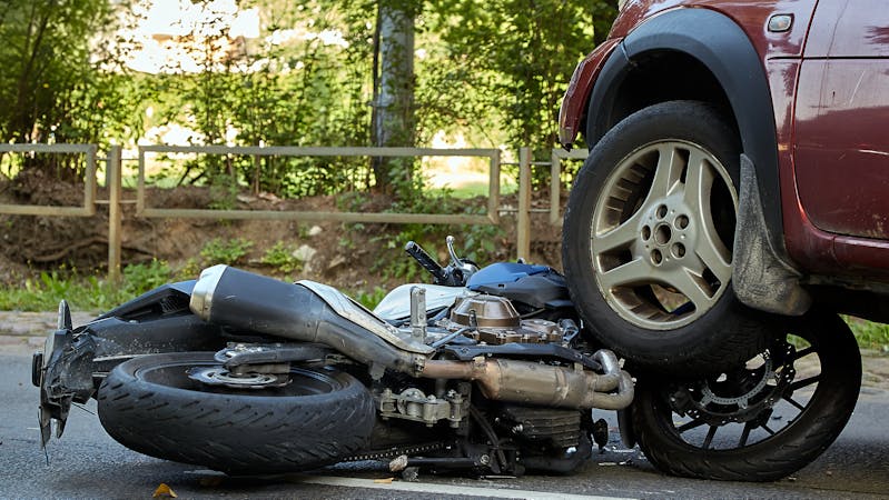 A motorcycle lies on its side, crushed beneath a car wheel. The scene is set on a road with greenery and a wooden fence in the background, indicating an accident.