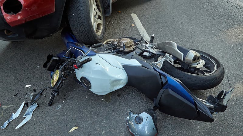 A damaged motorcycle lies on its side on the road, with debris scattered around it. The motorcycle is partially underneath a parked vehicle, indicating a collision or accident situation.