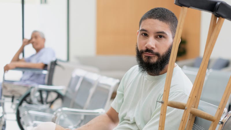 A man with a beard rests on crutches in a medical facility, with an older man in a wheelchair blurred in the background.