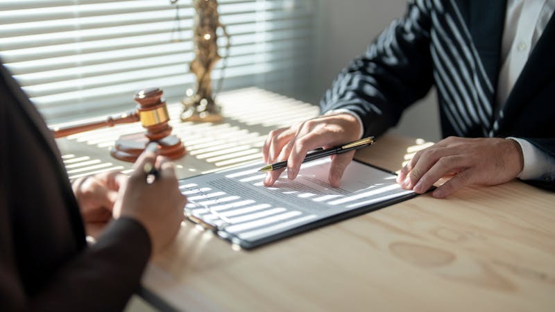 A document rests on a wooden table as one person points to it while another writes with a pen. Sunlight filters through blinds, casting shadows on the surface. A gavel is visible nearby.