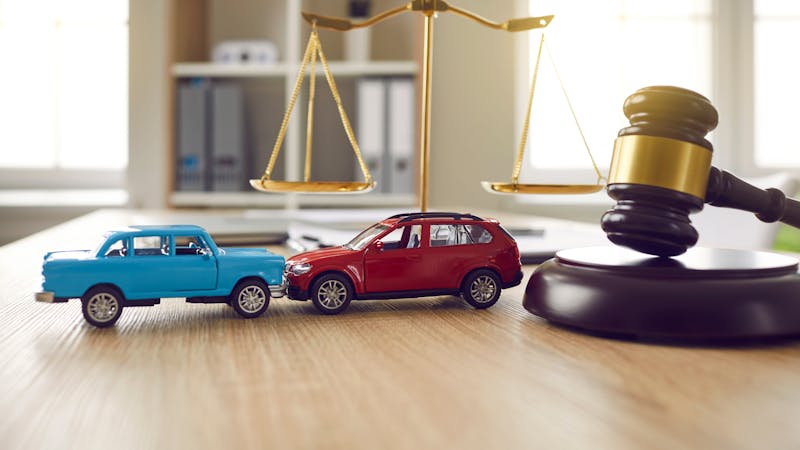 Two toy cars, one blue and one red, collide in close proximity to a golden balance scale and a black gavel, on a wooden surface in a well-lit room.