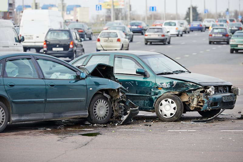 A damaged green car with its front end crumpled sits on a road with moving traffic in the background, indicating a recent collision.
