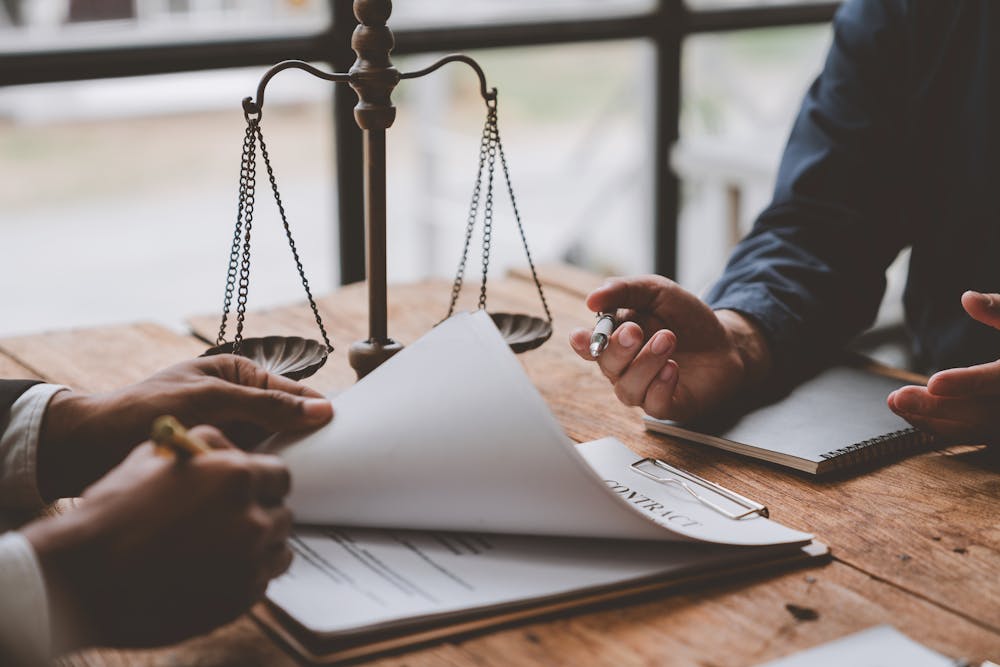 | Matz Injury Law Scales symbolize law atop a table where two people discuss a contract, one holding a pen, against a backdrop of large windows.