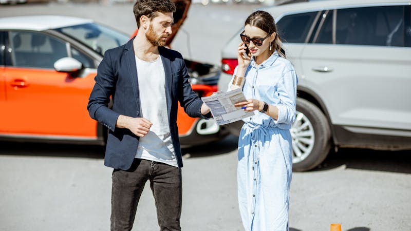 A man examines paperwork while a woman talks on the phone, standing next to two cars involved in a minor accident on a sunny day.