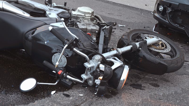 A damaged motorcycle lies on its side on a street, with a bent mirror and an exposed engine, surrounded by debris from an accident and a nearby car.