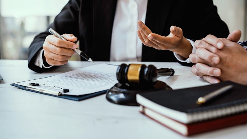 A person in a suit gestures while discussing documents on a table. A gavel rests nearby, and a second person's hands are visible, suggesting a legal consultation in an office environment.