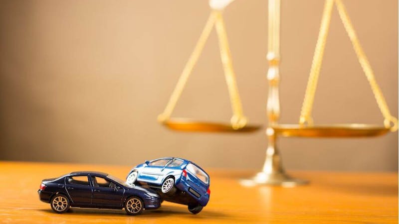 Two toy cars, one blue and one black, are positioned on a wooden surface, with the blue car atop the black one, while a balance scale stands in the background.