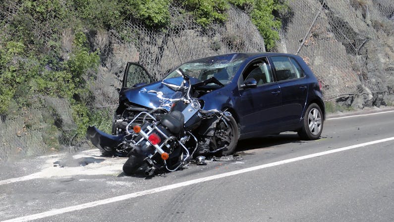 A damaged motorcycle lies on its side next to a blue car, which has also sustained significant front-end damage, on the roadside near vegetation and a rocky barrier.