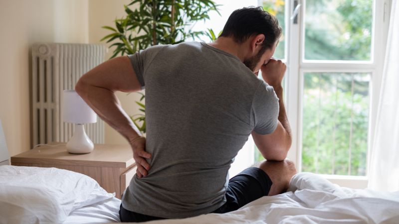 man in bed holding his back in pain on the edge of his bed