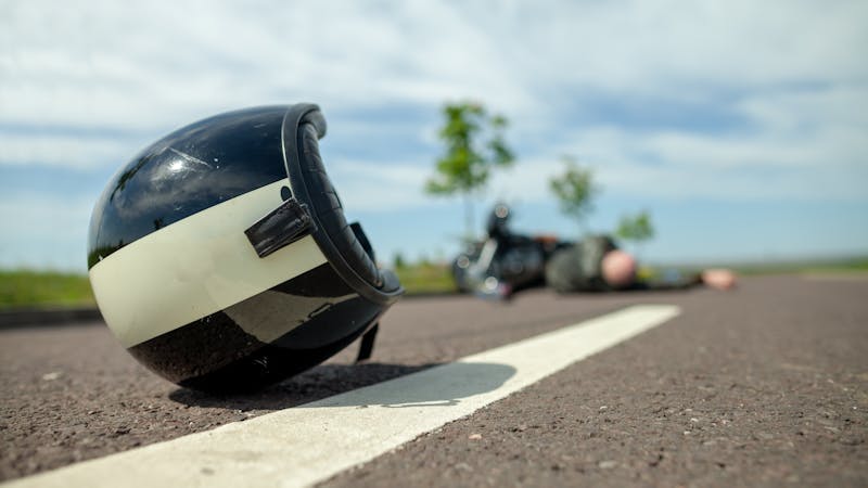 A black and white motorcycle helmet lies on the road, cracked, while a motorcycle and a person, possibly injured, are visible in the background under a clear blue sky with green trees nearby.
