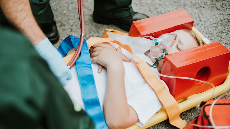 | Matz Injury Law A person receives medical attention, secured on a stretcher with vivid straps, oxygen mask on face, emergency responder nearby, suggesting a medical emergency or accident context.