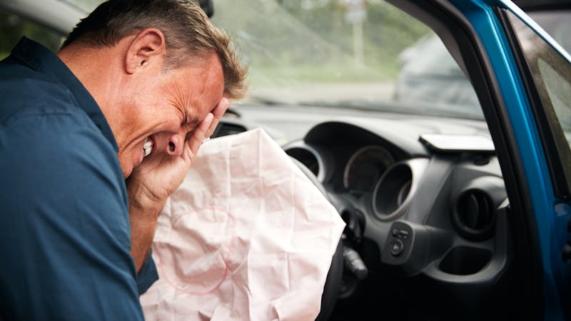 A man sits in a car, covering his face in distress, with an inflated airbag partially obstructing him. The interior is visible, with dashboard elements and a blurry external environment behind the windshield.