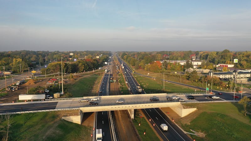 | Matz Injury Law A highway featuring multiple lanes is busy with vehicles moving in both directions. An overpass extends across the highway, with green trees and buildings lining the sides, under a clear sky.