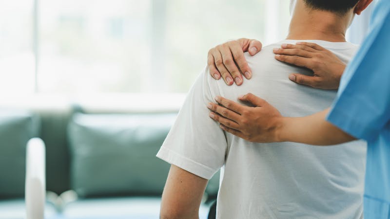 A healthcare professional is palpating a patient's shoulder in a clinical setting with a couch and windows in the background.