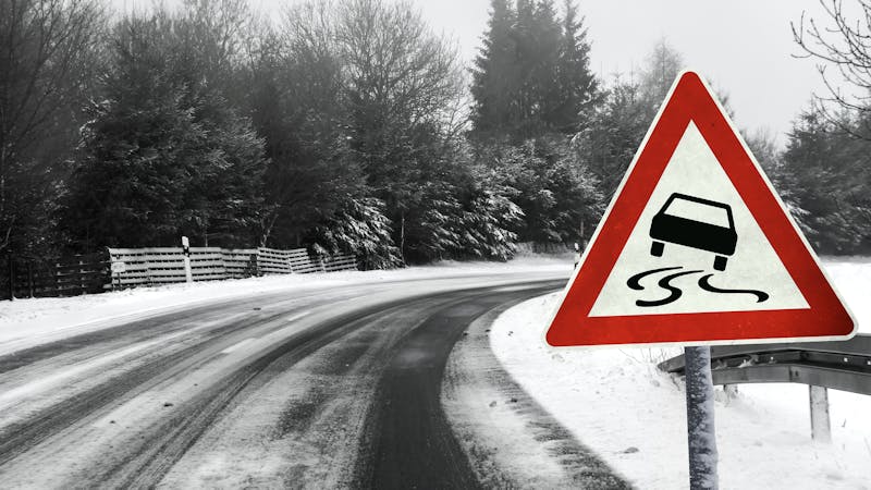 Slippery road warning sign on snowy winter road with trees in background.