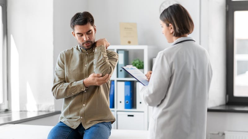 A man is sitting on an examination table, holding his arm and appearing concerned. A healthcare professional stands nearby, reviewing a clipboard in a modern medical office setting.