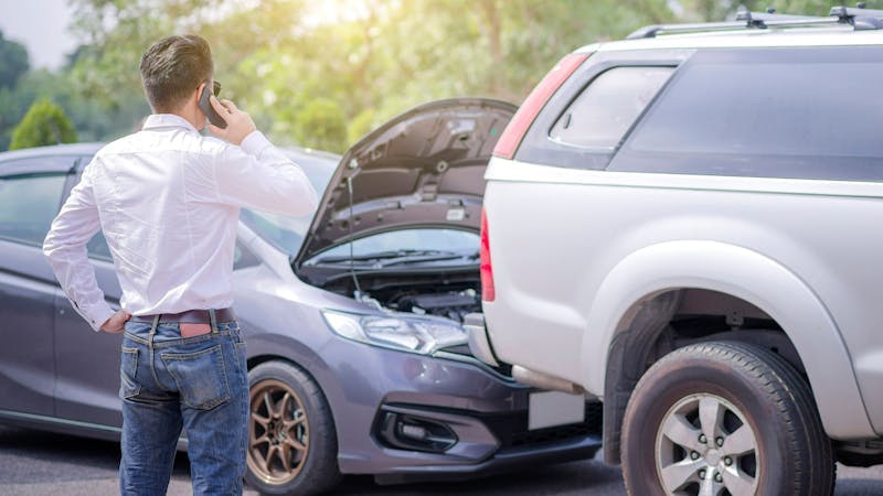 A man speaking on a cell phone stands beside two cars with open hoods, possibly dealing with a breakdown on a sunny roadside.