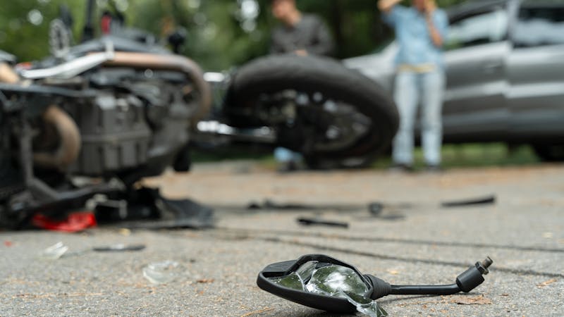 A shattered motorcycle mirror lies on the asphalt, with a fallen motorcycle nearby. Two individuals stand in the background, suggesting the aftermath of an accident in a wooded area.