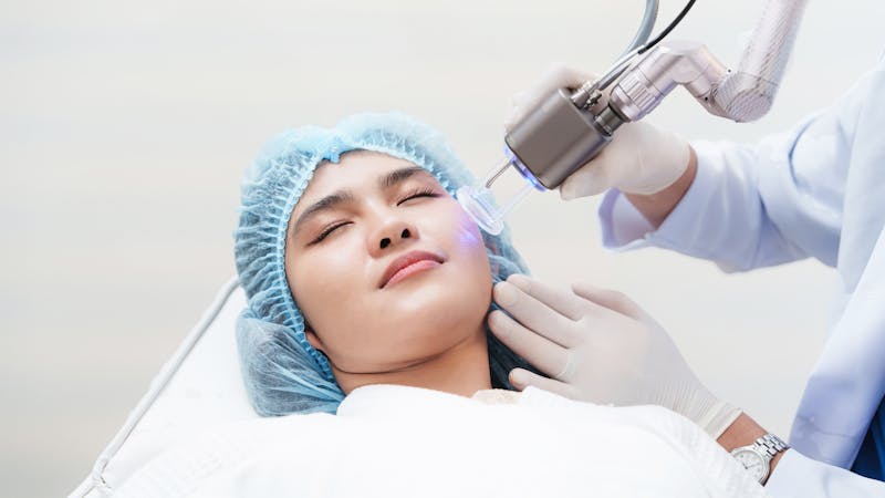 A person with a blue surgical cap relaxes in a reclined position while a professional, wearing gloves, uses a facial treatment device on their skin in a clinical setting.
