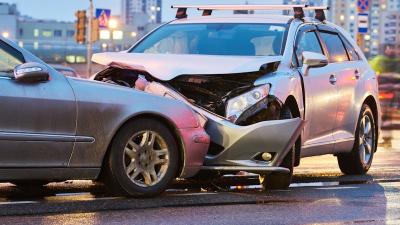 | Matz Injury Law Two cars are involved in a collision on a wet road. The silver car has significant front damage, while the gray car shows side damage. The background includes urban buildings and traffic signs.