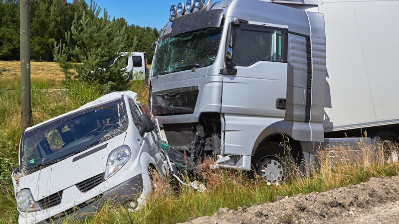 A white car is crashed into the front of a silver truck on the roadside, surrounded by grass and trees under a clear sky.