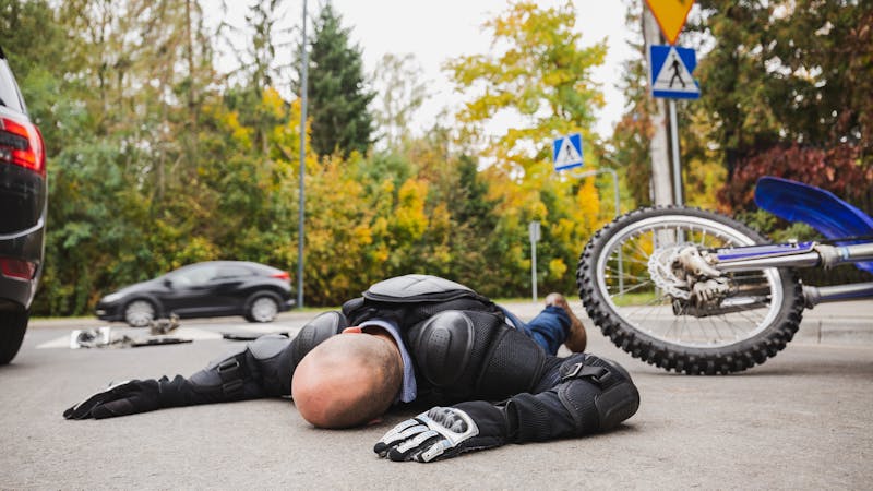 Motorcyclist in protective gear lying on road after accident, with bike fallen nearby and traffic signs in background.