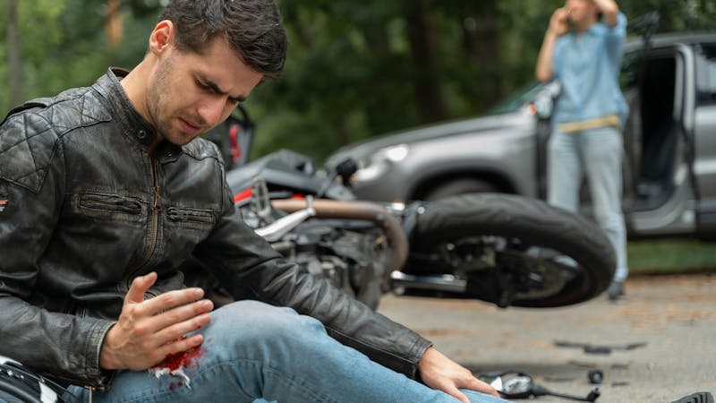 A man in a leather jacket sits on the ground, examining a bleeding injury on his hand. A motorcycle lies nearby, while another person is seen in the background using a phone.