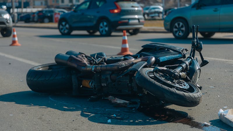 | Matz Injury Law A motorcycle lies on its side on a road, with visible damage and spilled fluid around it. Traffic cones are placed nearby, and cars are parked in the background.