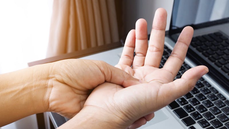 | Matz Injury Law A person's hands in focus, one pressing into the other's palm in a self-examination gesture, with a laptop and window blinds in the background.