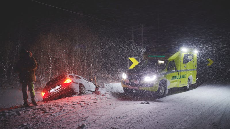 | Matz Injury Law A white car is partially buried in snow beside a dimly lit road, as an ambulance with flashing lights approaches. A person stands in the snow, observing the scene amidst a snowstorm.