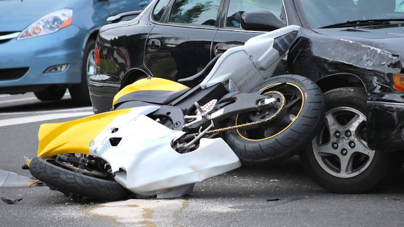 A yellow and white motorcycle is wrecked against a black car on a street, indicating a recent traffic collision.