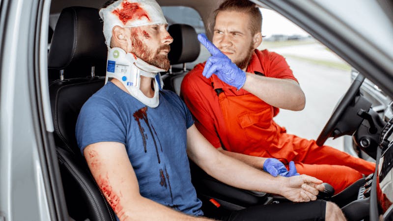 A bloodied man with a neck brace sits in the passenger seat of a car, while a paramedic, wearing blue gloves, urgently gestures and speaks to him in a concerned manner.