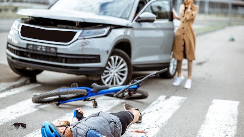 A cyclist lies on the ground, injured, near a fallen bike. A gray SUV is stopped nearby, and a woman in a coat is on the phone, appearing distressed.