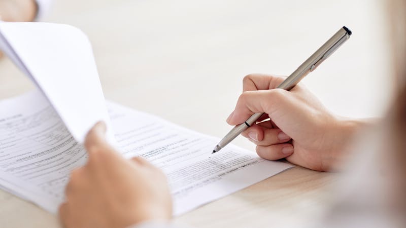 A person is signing a document on a desk, providing a close-up view of their hand holding a pen with text-filled papers beneath.