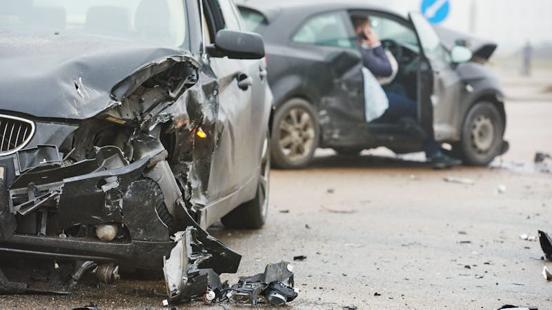 Two damaged cars after a collision, with debris scattered around on a street; a person stands in the background making a phone call.