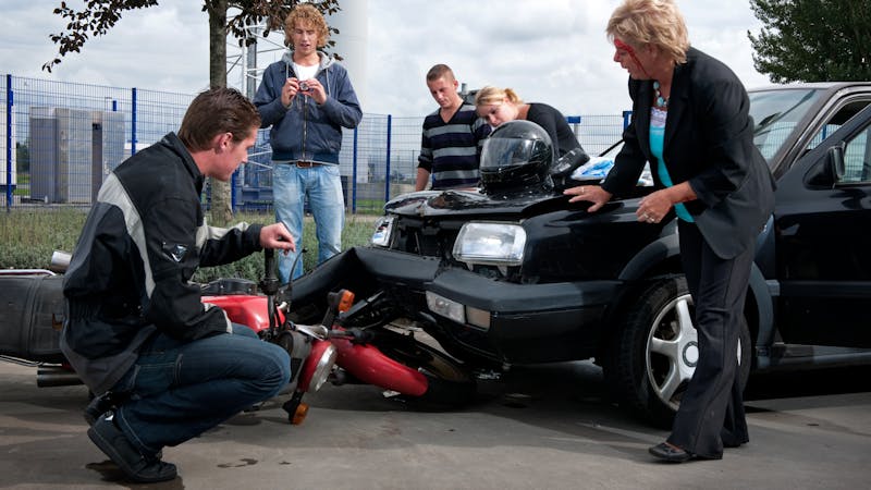 A group of people inspecting a car-motorcycle accident scene with a damaged front bumper on a cloudy day.